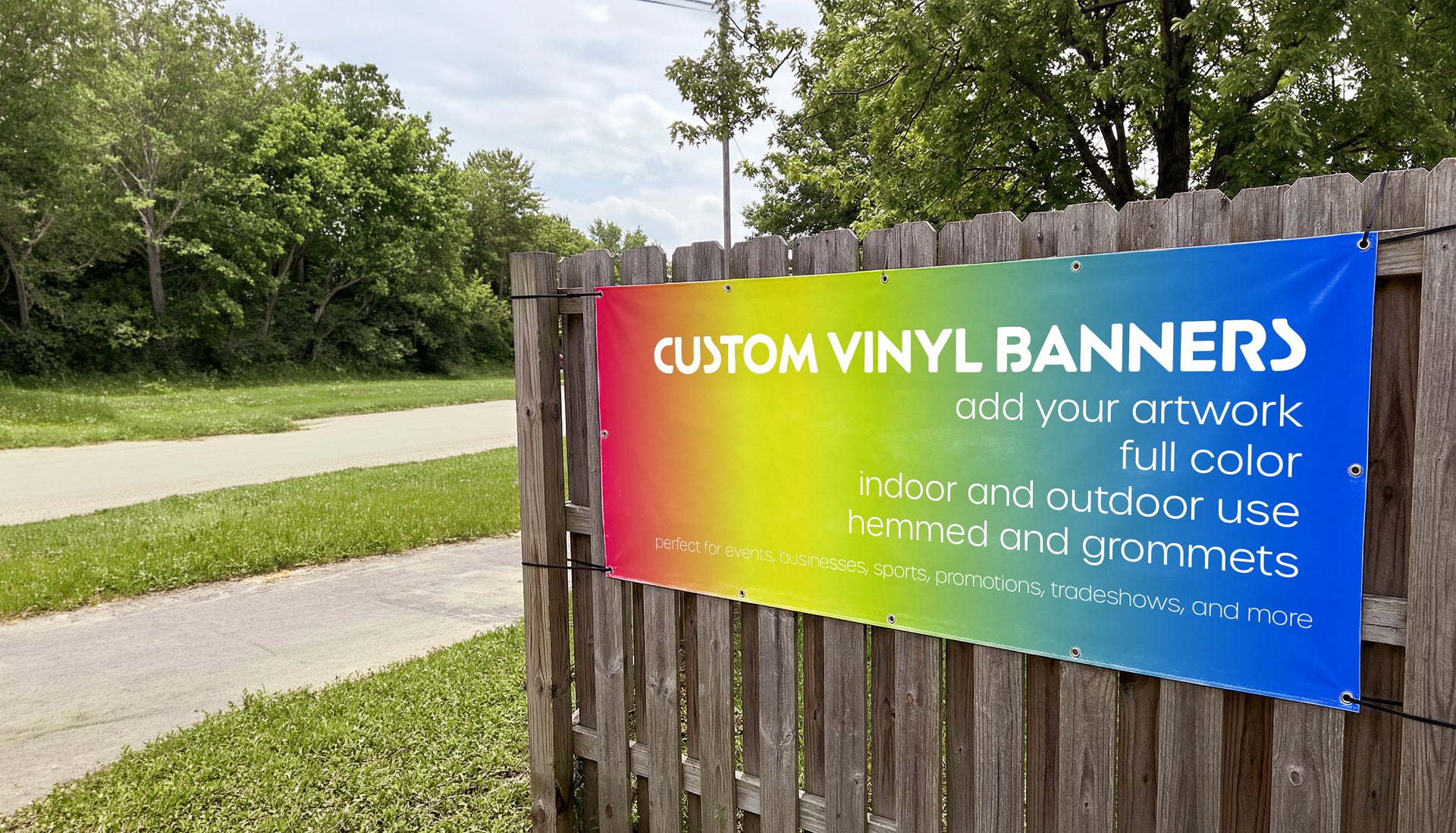 Colorful sign advertising custom vinyl banners on a wooden fence with trees in the background.