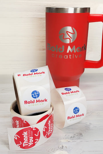 a red tumbler with a bold mark creative logo engraved on it and rolls of circle labels on a white background