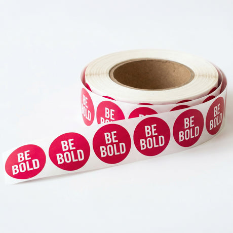 Roll of pink stickers with 'BE BOLD' text on a white background