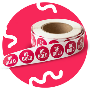 Roll of red and white 'BE BOLD' stickers on a pink background
