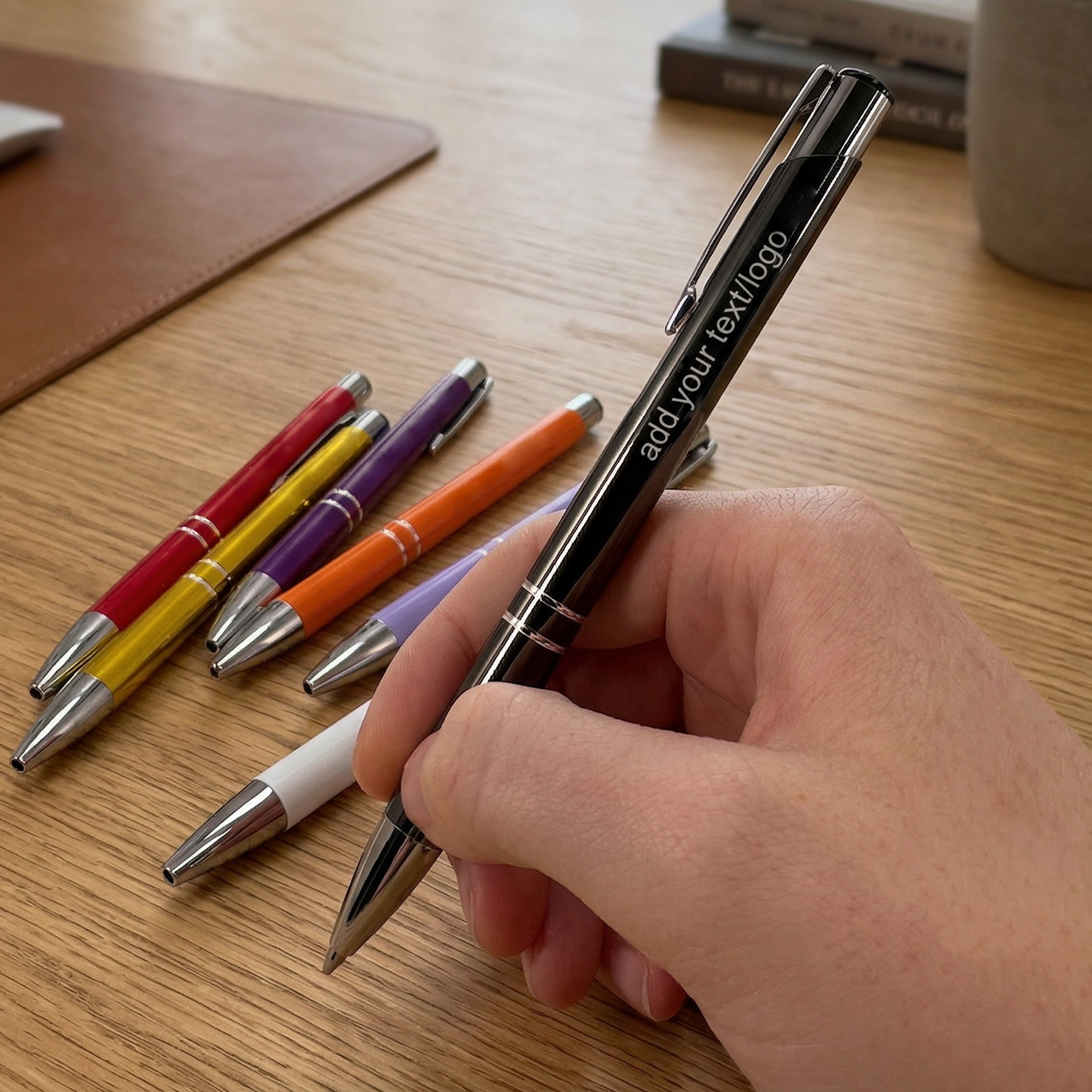 Hand holding a black pen with customizable text on a wooden surface with other colored pens.