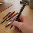 Hand holding a black pen with customizable text on a wooden surface with other colored pens.