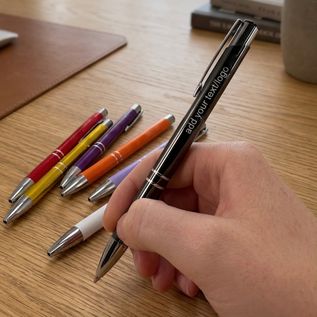 Hand holding a black pen with customizable text on a wooden surface with other colored pens.