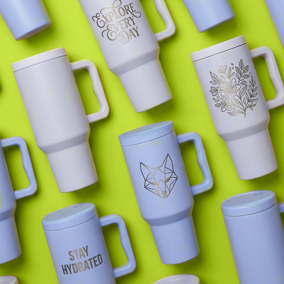 White and light blue travel mugs with various engraved designs on a green background