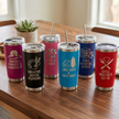 Set of six insulated tumblers with various engraved designs and text on a wooden table.